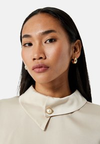 Un primer plano de una persona que lleva una blusa crema con un cuello redondeado y estructurado, y un botón dorado con un acento de perla. Se ven pendientes de aro dorados.