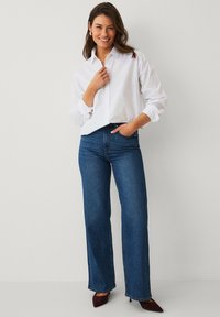 Chemise blanche à boutons avec manches retroussées, associée à un jean large bleu taille haute et des talons bordeaux à bouts pointus. Design simple, coupe classique.