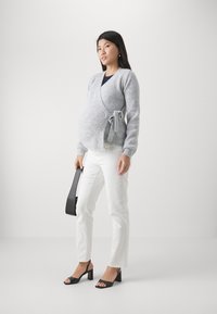 Cardigan enveloppant en tricot gris avec des manches longues, détail de lien à la taille, associé à un jean blanc et des sandales à talons carrés noirs.