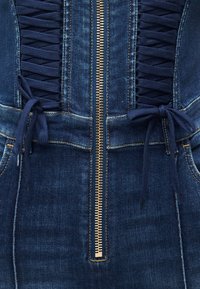 Κοντινό πλάνο σκούρου μπλε τζιν ένδυμα με κεντρικό χρυσό φερμουάρ και λεπτομέρειες δεσίματος με κορδόνια σε ναυτικό μπλε, δεμένα σε φιόγκους και στις δύο πλευρές.