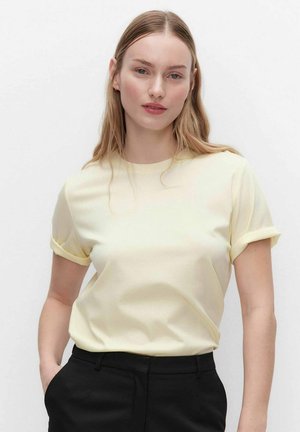 Jonge vrouw met lichtbruin haar draagt een lichtgele t-shirt met opgerolde mouwen, ingestopt in een zwarte broek, staand tegen een effen witte achtergrond.