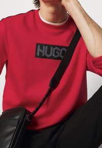 Felpa rossa con logo rettangolare nero "HUGO", con scollo a girocollo e accenti laterali. È visibile una borsa nera con tracolla intrecciata.