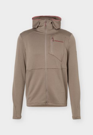 Chaqueta con capucha y cremallera color marrón claro, con tejido texturizado, bolsillo con cremallera en el pecho y logo rojo de "Peak Performance" en el pecho izquierdo.