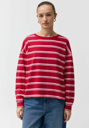 Kobieta ubrana w czerwoną koszulę z długim rękawem w białe poziome paski i niebieskie dżinsy, stojąca na jednolitym jasnym tle.