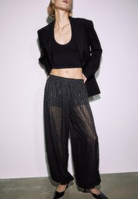 Crni skraćeni top bez rukava uparен s prozirnim, plisiranim crnim hlačama s blistavim detaljima i elastičnim pojasom. Crni blejzer zaokružuje izgled.