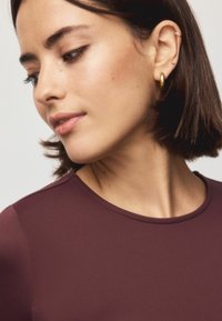 Bordeauxfarbenes Oberteil mit runder Ausschnitt, glatter Textur und taillierter Passform. Modell hat kurze braune Haare und goldene Creolen.