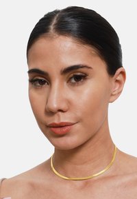 Zlatý kovový choker náhrdelník s hladkým, plochým dizajnom. Povrch má jemnú textúru a jednoduchý, elegantný tvar.
