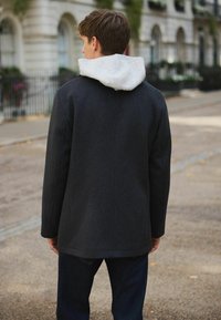 Young man wearing a black checked coat with gray hooded sweatshirt underneath, standing on a city street with blurred buildings in background.