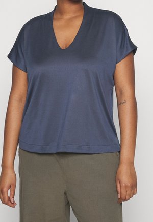 Personne portant un t-shirt bleu marine à manches courtes et col en V et un pantalon vert olive, montrant un petit tatouage sur l'avant-bras gauche.