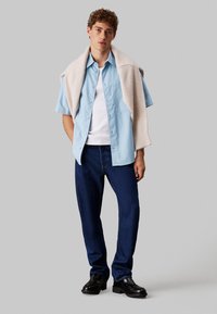 Camicia a maniche corte azzurro chiaro con bottoni indossata sopra una canottiera bianca, abbinata a jeans blu scuro e scarpe nere. Maglione appoggiato sulle spalle.