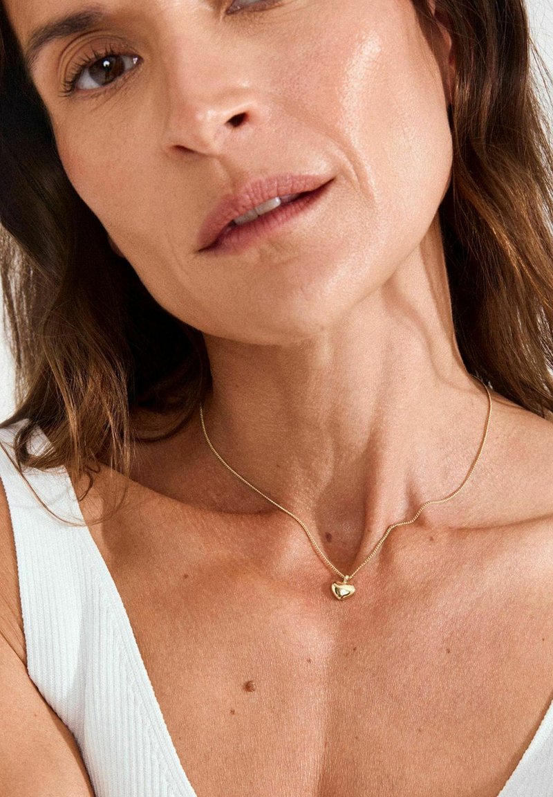 Gouden hartvormige hanger aan een fijne ketting, gedragen om de hals. De huid heeft een natuurlijke textuur en de achtergrond is neutraal en zacht verlicht.