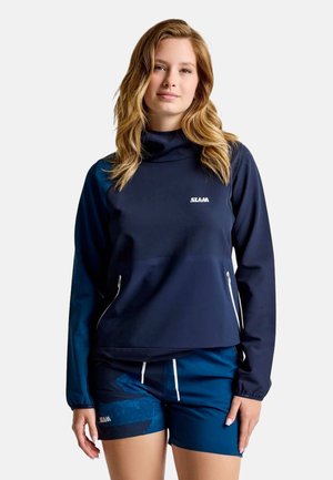 Frau mit langen, welligen Haaren, die einen marineblauen Langarm-Pullover mit Reißverschlusstaschen und passende marineblaue Shorts mit weißem Kordelzug vor schlichtem Hintergrund trägt.