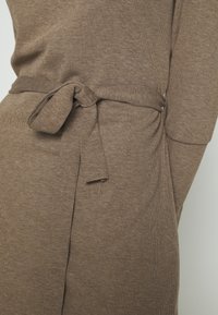Hněd dress z jemné ribované látky s páskem na zavázání v pase a bočním rozparkem. Má dlouhé rukávy a pohodlnou, měkkou strukturu.