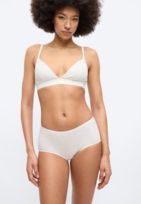 Γυναίκα με σγουρά μαλλιά που φοράει ανοιχτό γκρι σουτιέν και αντίστοιχο ψηλόμεσο εσώρουχο, στέκεται μπροστά σε απλό λευκό φόντο.