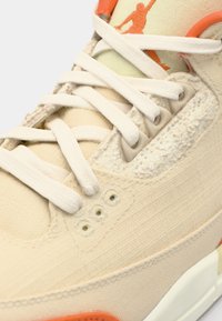 Zapatilla beige con una parte superior de tela texturizada, cordones blancos y acentos naranjas. Presenta una unidad de aire visible y detalles perforados en los laterales.