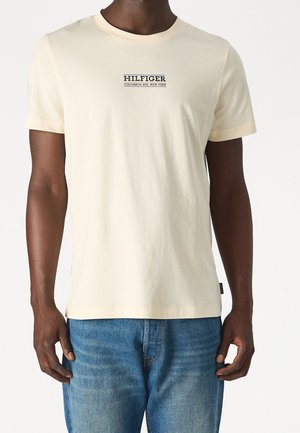 Homme portant un t-shirt crème uni à manches courtes avec le texte noir "Hilfiger Columbus Ave. New York" sur la poitrine et un jean bleu.