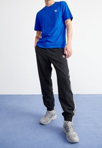 Blå bomulls-t-shirt med liten vit logotyp, kombinerad med svarta träningsbyxor med vita sidostreck och grå sneakers.