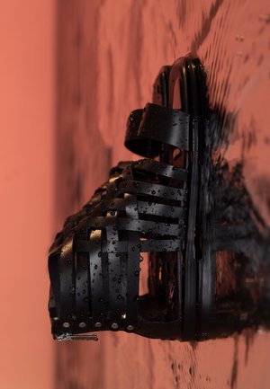 Sandalia negra de tiras con gotas de agua reposando sobre una superficie reflectante y húmeda contra un fondo naranja suave.