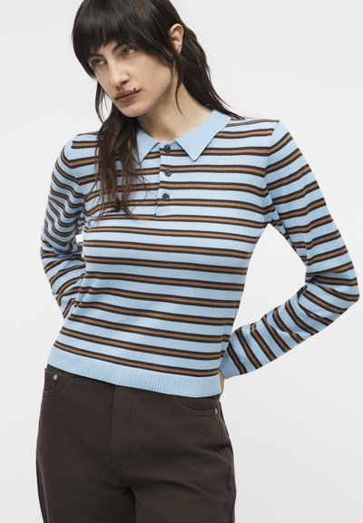 Jeune personne aux cheveux foncés portant un polo à manches longues rayé bleu clair et marron, ainsi qu'un pantalon foncé, debout les mains derrière le dos.