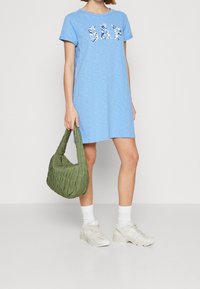 Robe T-shirt en coton bleu clair avec logo floral "GAP". Le mannequin tient un sac matelassé vert. Portée avec des chaussettes blanches et des baskets beige.