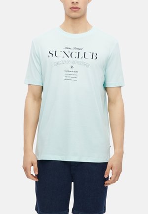 Mężczyzna noszący jasnoniebieską koszulkę z tekstem "Sunclub Ocean Sports" i ciemnoniebieskie spodenki, stojący na tle gładkiego jasnego tła.