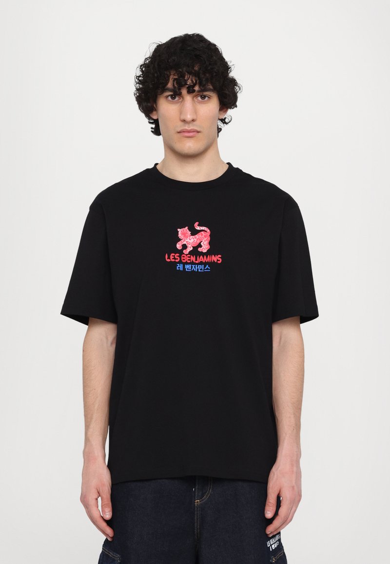 Un t-shirt en coton noir présente un graphisme rouge et bleu avec un lion stylisé et le texte "LES BENJAMINS" en caractères gras.