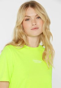 Neon green t-shirt with short sleeves, round neckline, and white text "Vivid Fusion SENSES.THE LABEL" on the front. Soft texture.