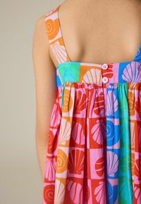 Fille portant une robe sans manches colorée avec des boutons roses, avec des motifs de coquillages orange, bleu, vert et rouge sur un fond clair.