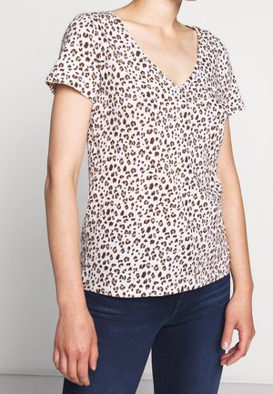 Vrouw draagt een top met korte mouwen en V-hals met luipaardprint, gecombineerd met donkerblauwe spijkerbroek, staand voor een effen lichte achtergrond.