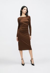 Καφέ μακρυμάνικο φόρεμα με ρυθμισμένες πιέτες, εφαρμοστή σιλουέτα και μήκος μέχρι το γόνατο, συνδυασμένο με μαύρα ψηλοτάκουνα παπούτσια.