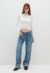 Weißes geripptes Crop-Top, hoch taillierte blaue Jeans mit umgeschlagenen Säumen und schwarze Knöchelstiefel. Das Model trägt getönte Sonnenbrillen und steht vor einem schlichten Hintergrund.