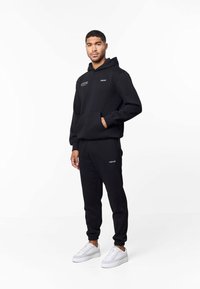 Man in een zwarte hoodie en joggingbroek met logo's, witte sneakers, hand in de hoodie-zak, tegen een witte achtergrond.