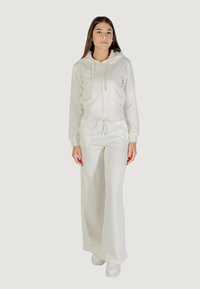 Set di tuta in pile bianco composto da una felpa con zip e cordini, e pantaloni larghi, entrambi con una finitura testurizzata. Le sneakers bianche completano il look.