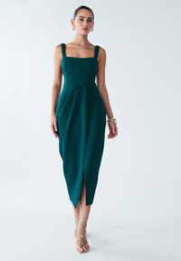 Turquoise midi-jurk met een vierkante halslijn, brede bandjes, een zijsplit en geplooid ontwerp. Gemaakt van textuurstof. Draagt bijpassende sandalen met bandjes.