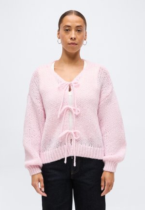 Kvinde med håret samlet bagpå iført store øreringe og en lyserød strikket cardigan med tre bindebånd foran over en hvid top.