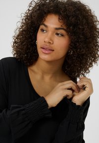 Top negro con un escote en V pronunciado y mangas fruncidas. Textura suave, con acentos de mangas abullonadas. El modelo se está arreglando el cabello.