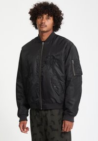 Giacca bomber nera in satin con polsini a coste, chiusura frontale con zip, tasche laterali e un distintivo decorativo nero sul petto.