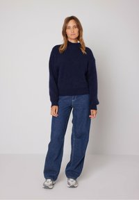 Tamsiai mėlynas megztinis megztas su aukštu kaklu ir laisvu kirpimu, derinamas su mėlynais džinsais ir pilkais sportbačiais.