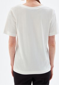 Bílý tričko s krátkým rukávem a kulatým výstřihem, hladká textura a volný střih, zobrazeno ze zadní strany, odhalující jednoduché švy.