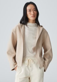Beige Zip-Up-Hoodie mit weicher Textur, weiten Ärmeln und angenähter Kapuze, getragen über einem hellen Crepehemd und hellen Cordhosen.