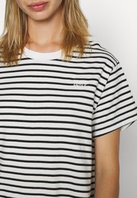 Musta-valkoraidallinen t-paita pyöreällä pääntiellä, lyhyillä hihoilla ja rinnan kohdalla brodeeratulla logolla. Kangas vaikuttaa pehmeältä ja rentoon käyttöön sopivalta.