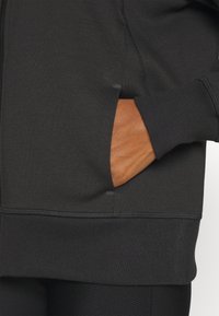 Svart zip-up-jacka tillverkad av ett slätt material, med en sidoficka och ribbade ärmar. Designen är minimalistisk utan synliga mönster.