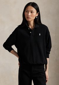 Naine, kellel on pikad mustad juuksed, kannab musta pikavarrukalist polo särki, millel on rinnal väike valge logo, seisab ühe käega puusal.