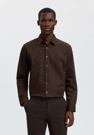 Homme debout face à l'avant portant une chemise marron foncé à boutons et un pantalon assorti, une main dans la poche.