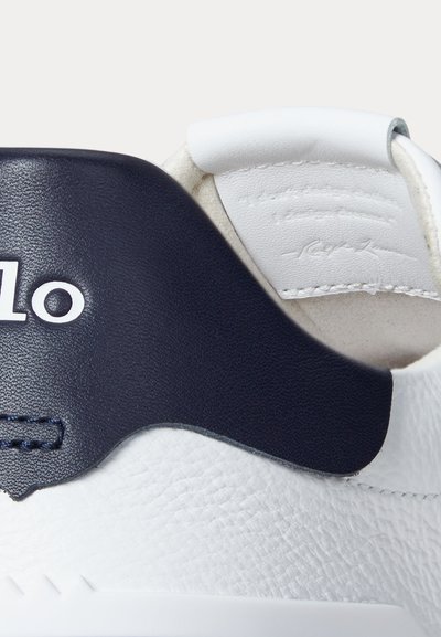 Tuvplānā balta ādas sporta kurpes papēdis ar tumši zilu detaļu un baltu polsterētu apkakli, ar daļēju baltu zīmola logotipu un izšūtām akcents detaļām.