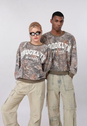 Twee modellen dragen bijpassende camouflage "Brooklyn New York" sweatshirts en beige cargobroeken, poserend tegen een effen lichte achtergrond.