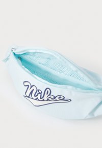 Šviesiai mėlynas liemenės paketas, pagamintas iš audinio, su akcentuota mėlyna Nike logotipu, užtrauktuko uždarymu ir tinkliniu kišeniu viduje organizavimui.