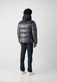 Mann iført en skinnende grå og svart puffjakke med hette, mørkeblå jeans, og hvite joggesko, stående med ryggen til mot en hvit bakgrunn.