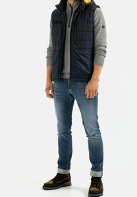 Námořnický prošívaný vest s kapucí přes šedou zipovou mikinu, modré džíny a hnědé-černé hikingové boty. Mezi výrazné prvky patří nadýchaná textura a akcenty.