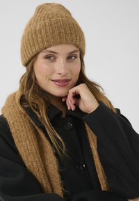 Bruine gebreide muts met ribbelstructuur, gedragen met een bijpassende sjaal. Zwarte jas met grote knopen, wat een gezellige winterlook benadrukt.